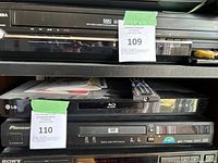 Photo of LG Blu-ray player with black remote on top, alongside other media players including a VHS player and a Sony CD player.