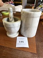 View of the white marble mortar and pestle next to the cylindrical marble wine cooler on a wooden surface, displaying natural veining and polished finish.