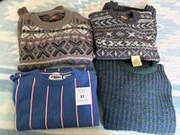 Four folded men's vintage sweaters on a light blue patterned background. Two patterned neutral tone sweaters on top; blue striped and green textured sweaters below.