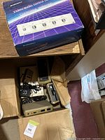 Open cardboard box containing assorted vintage electronics including Atari 5200 console and power adapters