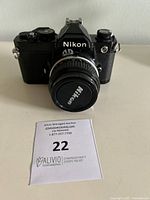 Front view of Nikon FM camera with attached lens and Nikon lens cap, showing the camera body and lens in black finish.