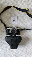 Olympus OM-1 film camera with attached F.Zuiko Auto-S 50mm lens, black leather case and camera strap laid on carpet.