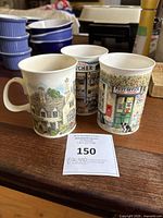 Three Dunoon fine bone china mugs displayed together, showing the detailed village storefront designs on each mug.