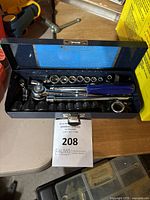 Open metal case showing ratchet handle with blue grip, multiple sockets laid out in recessed black plastic tray, and inside of lid with blue label