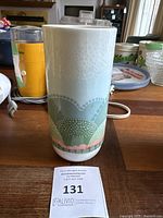 Photo showing tall ceramic vase with pastel Moj Dom pattern on wooden table indoors, auction tag '131' visible in front