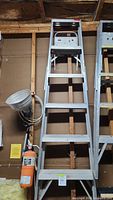 Full view of the folded aluminum step ladder leaning against wall. Shows ladder height and shelf.