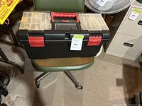 Black plastic toolbox resting on green chair showing red handle and latches with two removable clear plastic trays on top containing small parts.