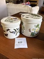 Two vintage ceramic canisters with lids showing floral and butterfly artwork on a wooden table.