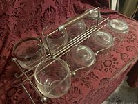 Six textured glass cups arranged in a metal rack with wooden handle on a patterned red fabric.