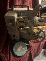 Gray 8mm film projector with metal reel and blue film reel on red patterned fabric.