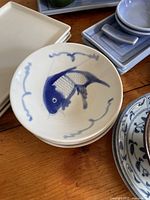 Close up of white shallow bowls with blue fish design and light blue square plates with condiment dishes in background.
