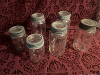 Six clear glass Crown jars with metal screw-on lids displayed on fabric background.