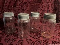 Four large clear glass Crown jars with metal screw lids standing on patterned cloth.