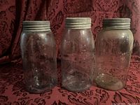 Three large Crown jars placed side by side on a dark red patterned fabric background, showing front embossed logos and metal lids.