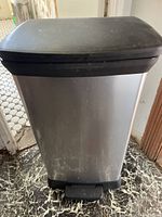 Front view of stainless steel rectangular step-on garbage can with black plastic lid and base on a marble floor.
