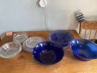 Clear and blue glass serving dishes set on wooden table against a plain wall, showcasing 9 pieces including 4 clear glass and 5 blue glass bowls/plates.