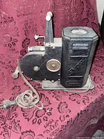 Side and front view of the hand crank 16mm film projector showing the crank handle, film housing, lens, and power cord on black textured metal body.