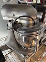 Front side view of vintage silver KitchenAid stand mixer with clear plastic pouring shield and attached stainless steel mixing bowl on metal rack.