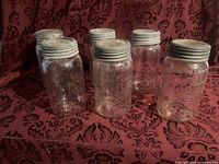 Six large clear glass jars with metal lids arranged on patterned red fabric background.