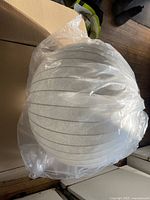 White spherical pendant light with ribbed black lines, wrapped in clear plastic, shown on top and inside cardboard box.