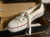 Side view of white canvas Converse sneaker inside original box, showing metal eyelets and laces, with red stripe on sole.