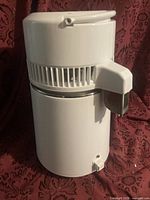 Photo of white water distiller machine on fabric background, showing spout and vented top section.