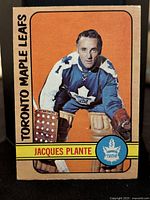 Front image of the hockey card showing Jacques Plante in goalie uniform for the Toronto Maple Leafs with orange background.