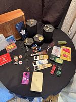Full assortment of vintage items displayed on black round table including jewelry, matchbooks, marbles, toy cars, pins, and small religious items