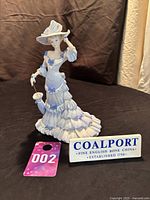 Full view of the Coalport figurine on black fabric background with label for identification and lot number 002 tag.