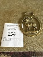Single metal horse brass ornament with raised stag or deer, circular scalloped frame, and handle at top. Surface shows wear and patina from age.
