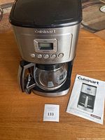 Front view of stainless steel Cuisinart coffee maker with digital display, buttons, glass carafe and user manual on table.