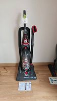 Full view of black and red upright Dirt Devil vacuum standing on wood floor with flexible hose attached and manual in front.