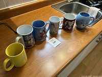Six assorted mugs on kitchen counter featuring Disney characters and a yellow Smokey Tunes mug