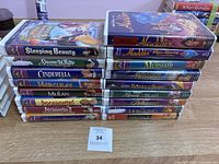 Photo showing the full collection of 15 Disney VHS tapes stacked and laid out, titles visible along the spine and front, including Sleeping Beauty, Snow White, Cinderella, Aladdin, Mulan, and others.