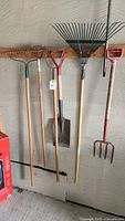 Full view of lot showing five garden tools hung on wall, including a garden rake, three-prong cultivator, shovel, leaf rake, and garden fork with red handle.