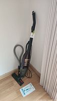 Full upright vacuum cleaner with hose and cord, positioned in corner on hardwood floor with manual beside it