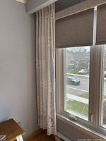 Curtain hanging on a window with beige roller shades, wooden furniture beside it, showing full length and floral lace pattern.