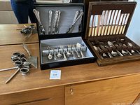 Overview of flatware and cutlery sets arranged on wooden surface showing black box with silver utensils, wooden box with beige handled knives, and loose silver-plated items.