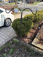 Black metal outdoor plant hanger with two hooks, placed outdoors next to garden and driveway.