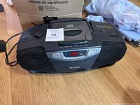 Front and top view of the black Venturer radio with CD player, showing the control panel, display, and speaker grills, placed on a wooden floor with some clothing and a cardboard box nearby.