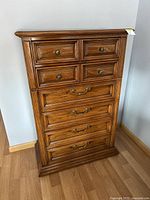 Front view of solid wood tall boy chest of drawers showing detailed wood paneling and brass handles on seven drawers.