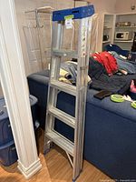 Front angle view showing four-step aluminum step ladder with blue top platform and rubber feet standing indoors near a white pillar and blue sofa with household items in the background.