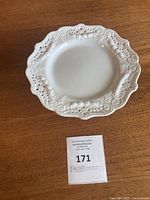 Top-down view showing ornate raised floral and scroll embossed design around scalloped rim of the white ceramic serving plate.