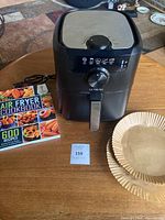 Front and slightly angled view of black Ultrean air fryer, showing dial and control icons on front panel, placed on wooden surface with cookbook and parchment liners next to it.