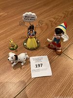 Four Disney character figurines on wooden floor: Snow White with sign, Pinocchio, Dalmatian puppy, and frog