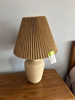 Full view of the vintage taupe beige ceramic table lamp featuring a pleated lampshade on a wooden surface against a plain wall.