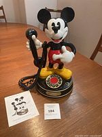 Front view of vintage Mickey Mouse talking telephone with handset held by the figure. Rotary dial visible on base with red center button. Instruction manual and lot tag placed beside the telephone.