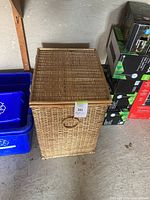 Photo of closed rectangular wicker laundry basket with lid and circular handle on front, placed by recycling bins and boxes