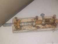 Photo of antique metal bolt lock showing heavy rust and worn white paint on rectangular base with sliding bolt and latch.