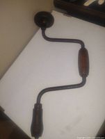 Antique hand brace wood drilling tool shown on flat surface, displaying curved metal shaft with wooden handles and central wooden grip.